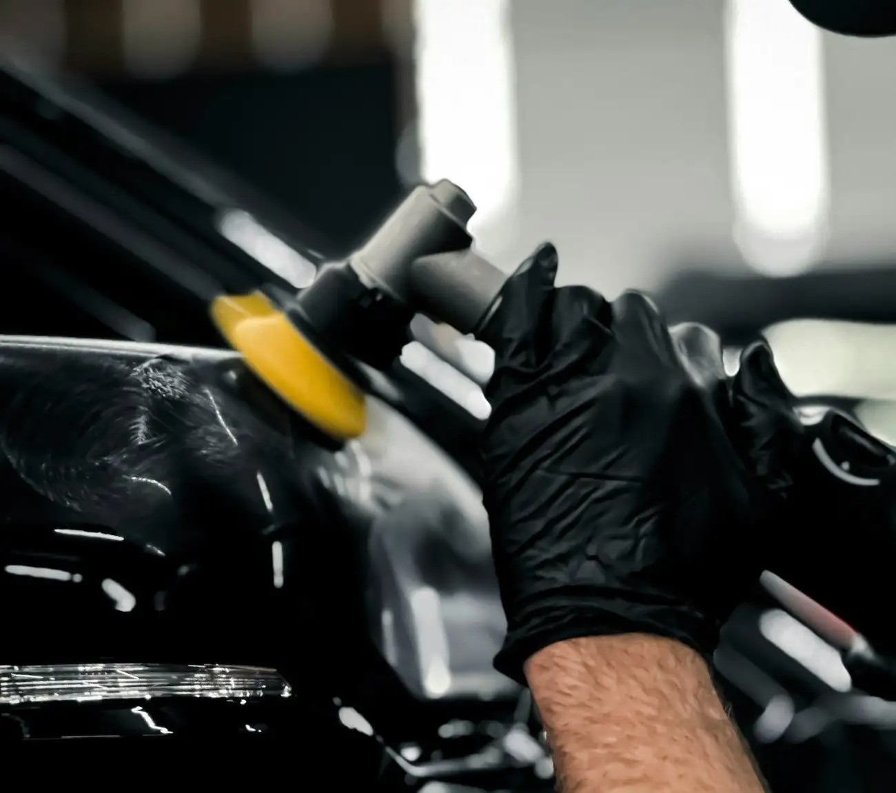 Gloved hand guiding a small yellow polishing pad along the edge of a black panel