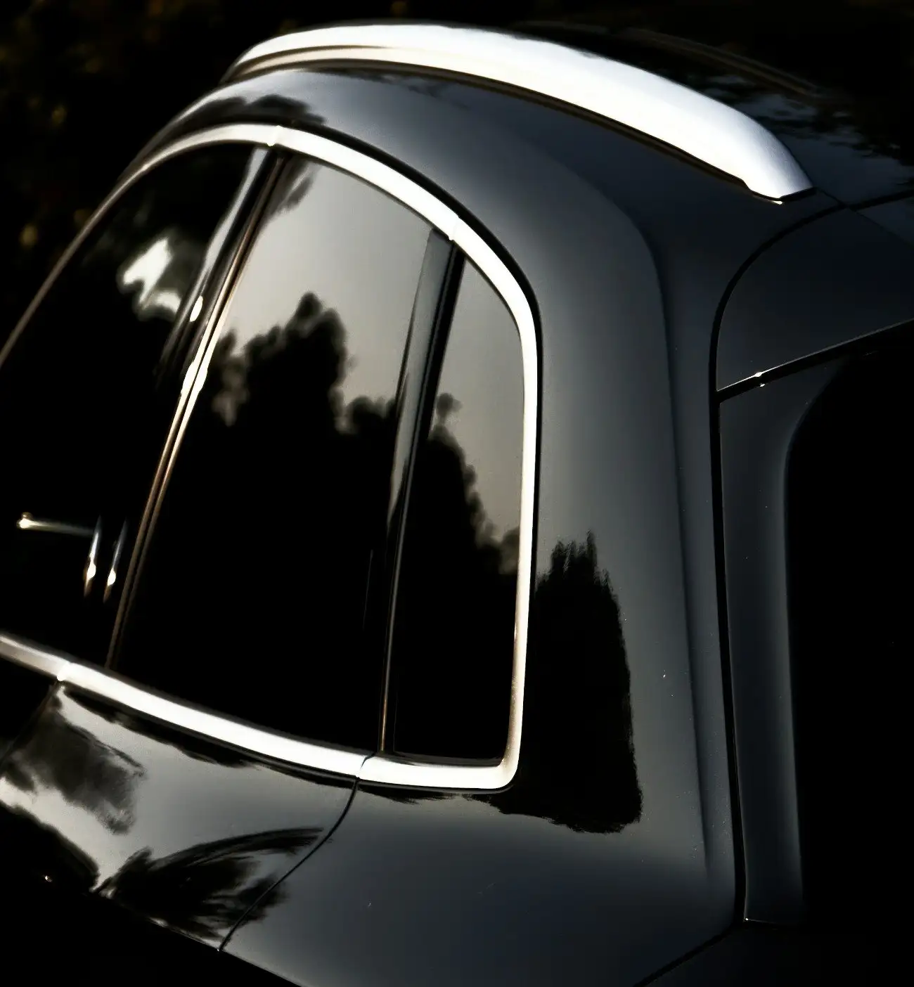 Rear side window and roof pillar of a black SUV with dark tinted glass reflecting trees