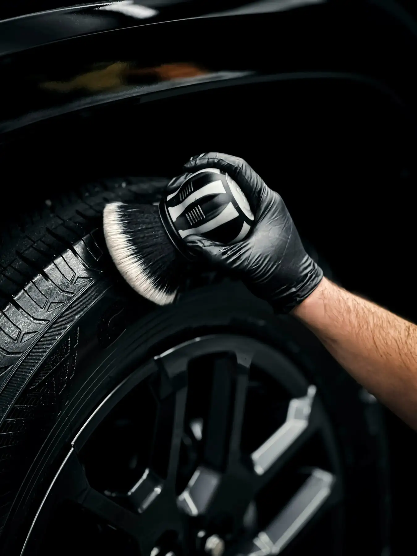 Gloved hand scrubbing a truck tire sidewall with a soft wheel brush, black wheel below