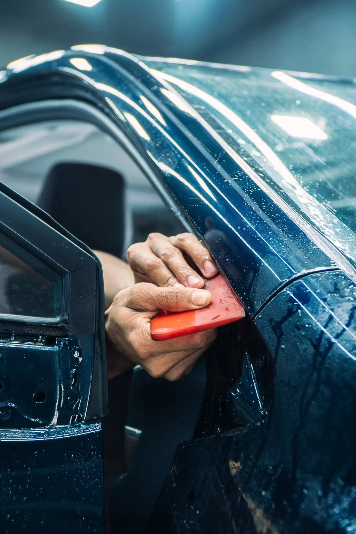 Installer's hands pressing clear film into the edge of a dark blue door with a red squeegee