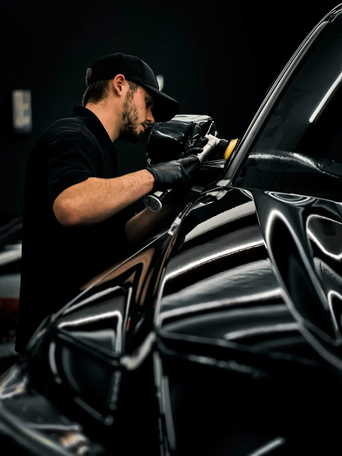 Detailer in black gloves machine-polishing the mirror housing of a black car under studio lights