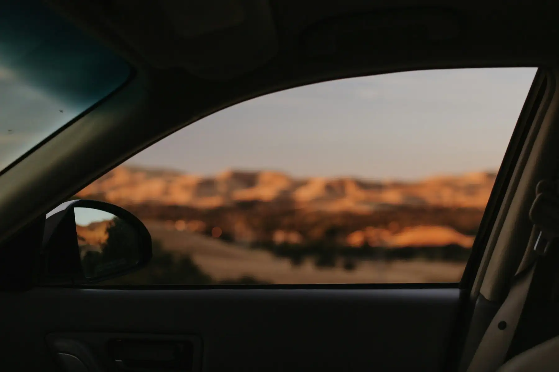 View from the driver's seat through the side window towards dry desert hills at dusk, the door mirror in the lower corner